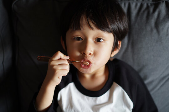 Little Asian boy, brushing his teeth on the sofa 