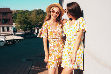 Two young beautiful smiling hipster female in trendy summer dresses  clothes. Sexy carefree women posing in the street. Positive models having fun, talking, chatting. Going crazy. In sunglasses