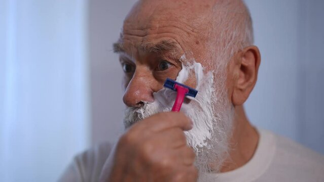 Headshot of gray-haired old Caucasian man shaving with razor in slow motion indoors. Focused senior retiree styling white beard in bathroom at home indoors. Lifestyle and hygiene concept
