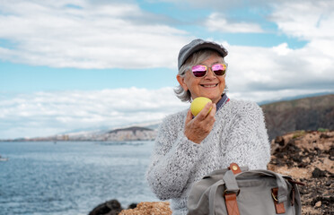 Fototapeta Happy handsome senior woman sitting by the sea eating an apple. Smiling elderly lady in outdoor excursion enjoying freedom, travel and retirement obraz