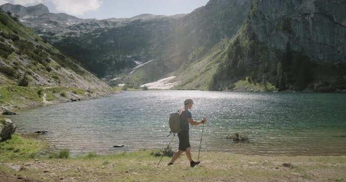Side tracking of a hiker walking with hiking poles near a mountain lake, water in the lake is glistening, mountains in the back.