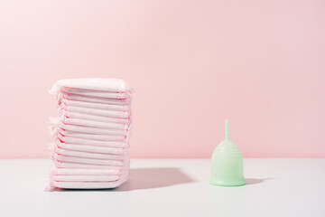 Menstrual cup next to pack of pads on white table against pink background.