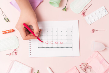 Lady marks with red felt-tip pen in the calendar the menstrual cycle on pink background with pills and sanitary napkin.