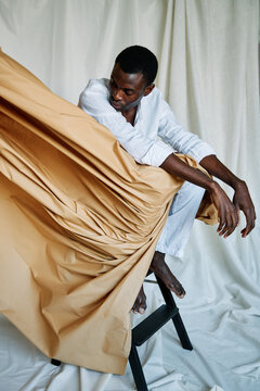 Black man sitting on white backdrop with a colour cloth.