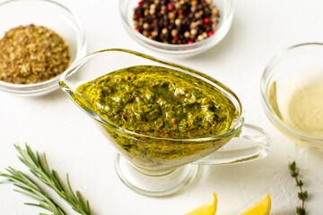 Chimichurri sauce in a gravy bowl on a white background. Various spices lie nearby. Argentinean vegetarian sauce made from olive oil, oregano, parsley.