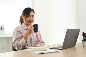 Pleased asian female employee drinking coffee and reading business email or online information on laptop computer