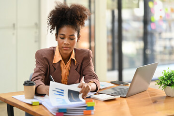 Concentrated African American female economist analyzing financial data and using laptop computer at work station