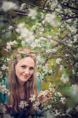 Fototapeta premium A cute girl with long blond hair in a jacket stands among flowering trees and smiles in spring.