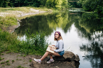 A young schoolgirl with stylishly dyed hair, with a purple lock of hair, in denim shorts and a white shirt, sits on a stone near a picturesque river with a big book in her hands.