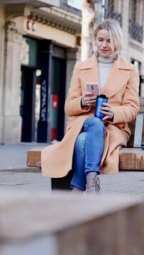 Stylish business professional blond woman checking cell phone sitting on street bench in winter