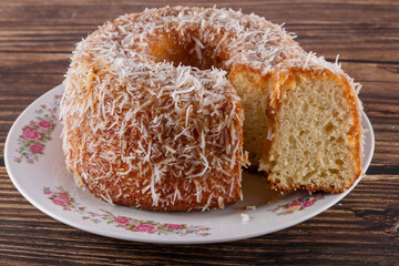 Coconut-flavored sweet cake topped with coconut flakes, with a cut slice showing the delicious dough on a decorated plate. Using wooden board and wooden background. Selective focus.