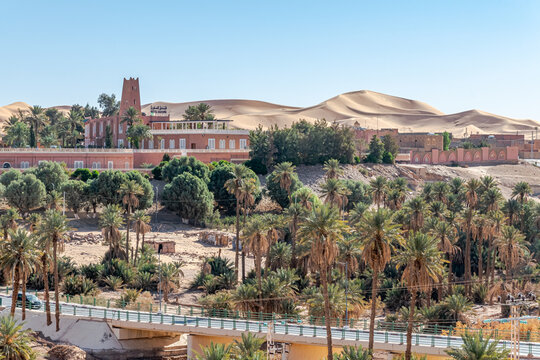 Taghit town of Bechar province in Algeria Sahara desert. Palm trees oasis, sand dunes and buildings. Road bridge of Oued Zouzfana river and Saoura hotel with an Algerian flag on the roof.