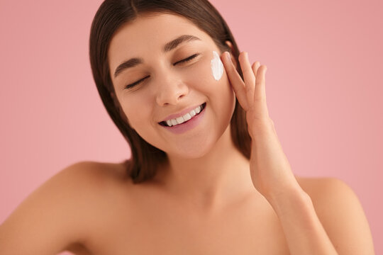Delighted young woman applying cosmetics on face and smiling in pink studio
