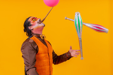 Portrait of a juggler in a vest with a painted face juggling with maces on a yellow background - obrazy, fototapety, plakaty