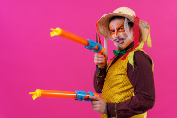 Clown with white facial makeup on a pink background, playing with toy guns and hat