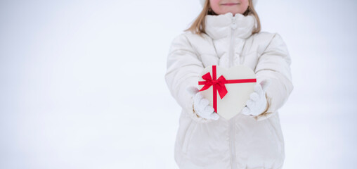 A joyful little girl in white winter clothes stands in front of the snow and holds a heart-shaped box