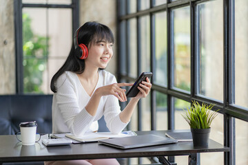 Young woman sitting at table wearing wireless headphones holding smartphone using video call greeting friend or client start talking via video conference