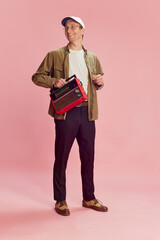 Cheerful mood. Young positive man in shirt, trousers and cap posing with vintage radio set over pink studio background. Music. Concept of emotions, facial expression, lifestyle, retro fashion. Ad