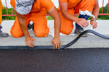 Workers apply black bitumen membrane strips for waterproofing roads and bridges to the curb of the...