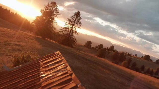 Aerial footage from FPV racing drone of a sun lit meadow during a summer sunset. Flying above an old barn and in between spruce trees. Sun is peaking through clouds creating beautiful lens flare.
