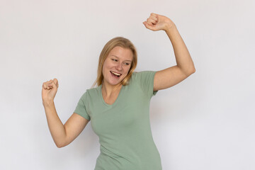 Fototapeta na wymiar Excited young woman dancing. Portrait of happy Caucasian female model with fair hair in green T-shirt looking away with raised hands, smiling, celebrating success. Victory, success concept