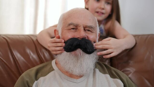 Front view close-up of positive senior bearded man with blurred girl placing toy black mustache on face of retiree. Relaxed Caucasian grandfather on weekend with playful granddaughter. Slow motion