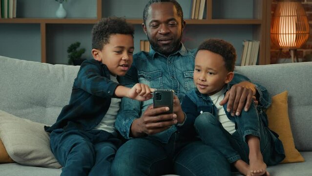 African American ethnic father man dad teach little boys brothers siblings children using mobile phone sitting on sofa at home daddy with two kids talking sit at couch shopping online with smartphone