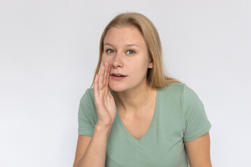 Fototapeta premium Secretive young woman whispering. Portrait of pleased Caucasian female model with fair hair in green T-shirt looking at camera, telling secret with hand near mouth. Gossip, announcement concept