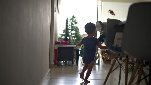 Slow motion of a latin baby boy running and chasing bubbles made by his father on a floor fan inside his home