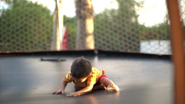 Slow motion of a latin toddler wearing a red short and a yellow shirt playing and laughing on a trampoline on a sunny summer afternoon