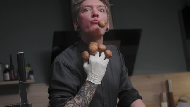 Young crazy male chef in an elegant black shirt with an alternative look, tattoos holding mushrooms, spitting one out.
A modern, fancy looking kitchen with fresh vegetables besides Cinematic