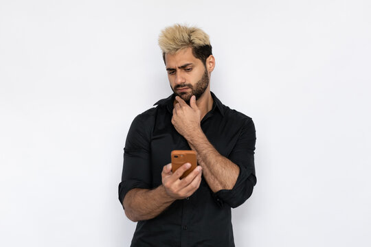 Hesitated young man reading news on smartphone. Male Caucasian model with brown eyes, ombre painted hair and beard in black shirt touching his chin reading message. Modern technology concept