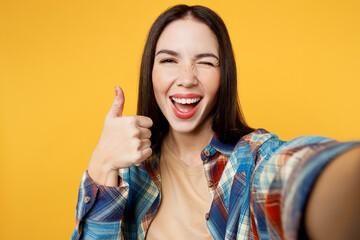 Close up young smiling woman wear blue shirt beige t-shirt doing selfie shot pov on mobile cell phone show thumb up wink isolated on plain yellow background studio portrait. People lifestyle concept.