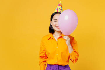Happy fun cheerful smiling young woman wear casual clothes hat celebrating covering half of face with pink balloon look camera isolated on plain yellow background. Birthday 8 14 holiday party concept