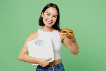 Young smiling cheerful fun woman wearing white clothes eat burger hold scales look camera isolated...