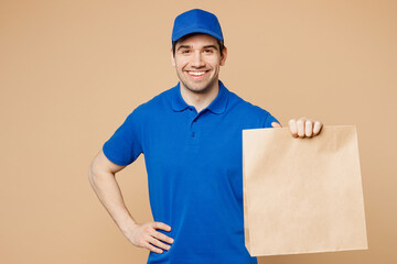 Fototapeta na wymiar Delivery guy employee man wear blue cap t-shirt uniform workwear work as dealer courier hold brown clear blank craft paper takeaway bag mockup isolated on plain light beige background Service concept