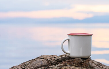 An enamel mug standing still on the top of a rock while showing the out of focus scenery at the...