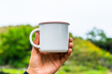 A hand holding an enamel mug while showing the out of focus scenery at the background