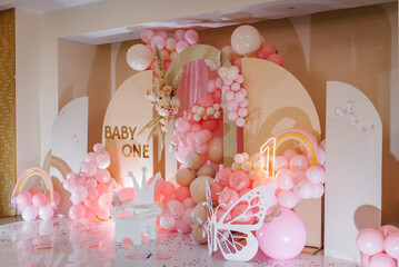 Birthday party for 1 year old girl on a background photo wall. Arch decorated pink balloons, rainbow, text baby one, flowers, paper decor butterfly, and wooden white chair. Children's photo zone.