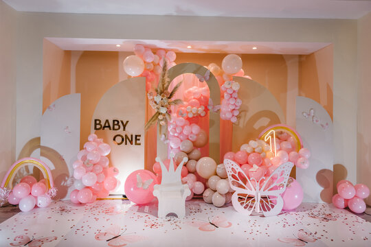 Children's photo zone. Birthday party for 1 year old girl on a background photo wall. Arch decorated pink balloons, rainbow, text baby one, flowers, paper decor butterfly, and wooden white chair.