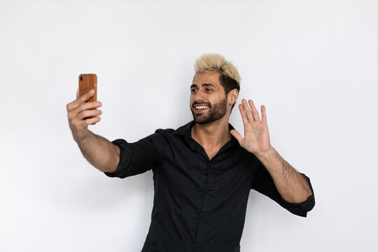 Joyful young man taking selfie. Male Caucasian model with brown eyes, ombre painted hair and beard in black shirt smiling taking self-portrait, waving his hand. Self-assurance, lifestyle concept