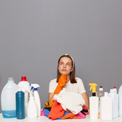 Photo of pensive Caucasian woman wears rubber protective gloves and headband, surrounded with bottles of detergents for laundry, looking up at copy space for advertisement.