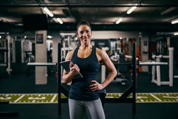 Poster A female personal trainer is posing in a gym with tablet in her hands and smiling at the camera. © dusanpetkovic1