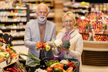 A healthy happy senior couple is holding fresh vegetables in hands at the supermarket while smiling at the camera.