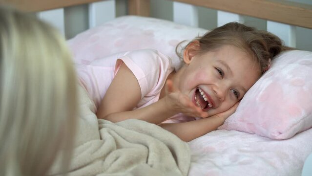 Back view of mother sits next to daughter in bed and puts sleep. Child laughs and naughty before bed. Healthy sleep concept