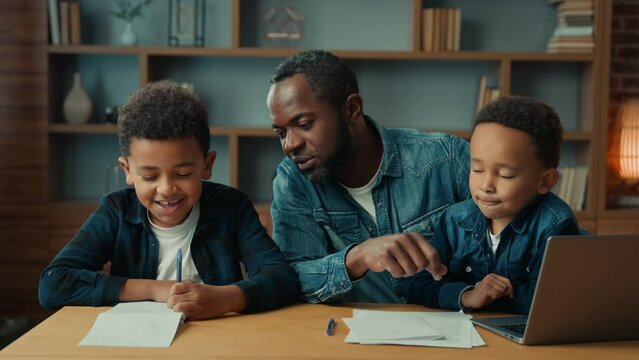 African American father dad teach schoolboy son help with homework sitting at table. Dad with two little boys children kids with laptop at home helping pupil write lesson class fatherhood childcare