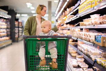 Mother pushing shopping cart with her infant baby boy child down department aisle in supermarket...