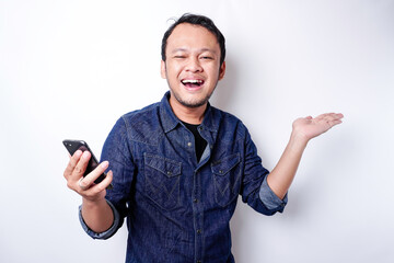 Excited Asian man wearing blue shirt pointing at the copy space beside him while holding his phone, isolated by white background