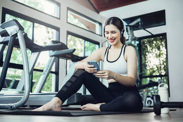 Fototapeta na wymiar Happy young woman wearing wireless headset and listening to music while resting, sitting on fitness mat after exercising in gym indoors.