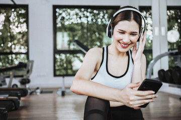 Happy young woman wearing wireless headset and listening to music while resting, sitting on fitness mat after exercising in gym indoors.